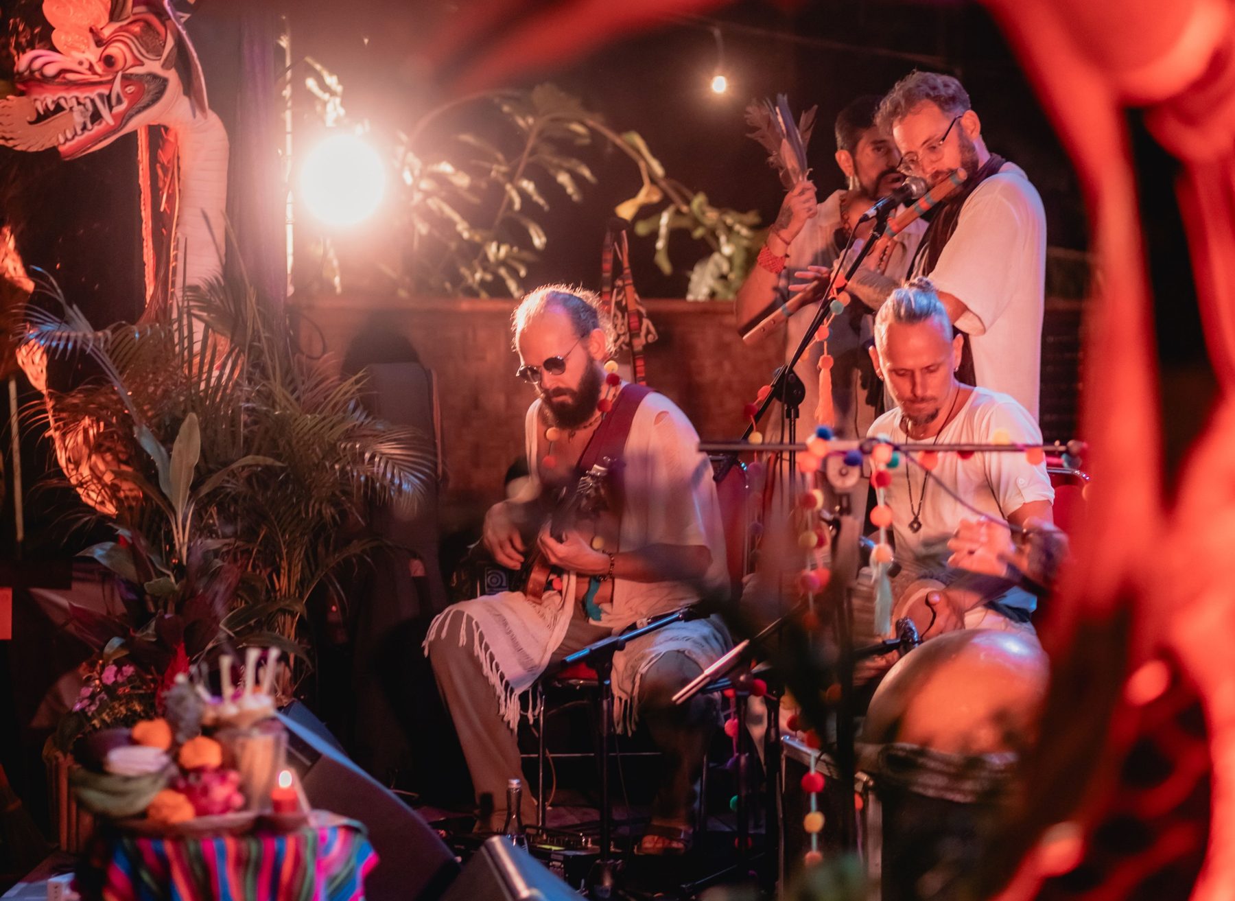 Acharya playing bass guitar with flute and percussion on a candlelit, flower-decked stage